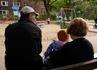 Rent an Oma Ein älterer Mann und eine ältere Frau sitzen mit einem kleinen Kind auf einer Bank auf einem Spielplatz. Sie sind nur von hinten zu sehen.