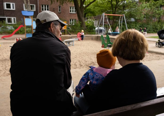 Ein älterer Mann und eine ältere Frau sitzen mit einem kleinen Kind auf einer Bank auf einem Spielplatz. Sie sind nur von hinten zu sehen.