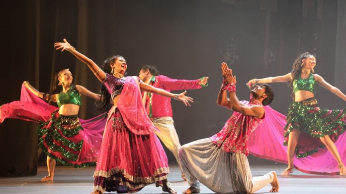 Frauen und Männer haben Indische Gewänder an und tanzen auf einer Bühne. Sie Tanzen einen traditionellen Indischen Tanz.