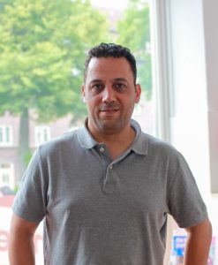 Ein Mann mit dunklen Haaren und einem grauen kurzärmeligen Polo-Shirt steht vor einem Fenster.