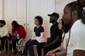 Workshop Teilnehmende sitzen auf Stühlen und lauschen interessiert den Erklärungen von Fayim.