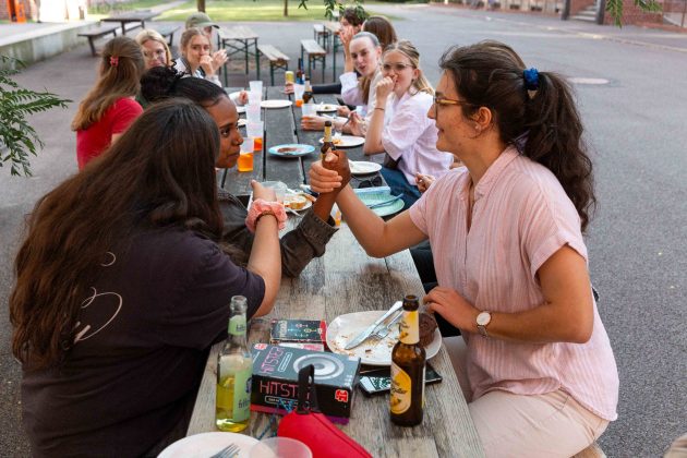 An einem langen Tisch sitzen mehrere Personen. Zwei Frauen spielen Armdrücken.