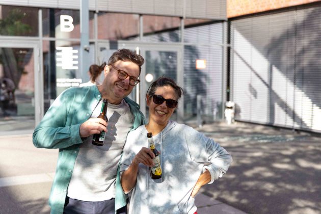 Ein Mann und eine Frau stehen nebeneinander, sie halten beide ein Getränk in der Hand und lächeln in die Kamera.