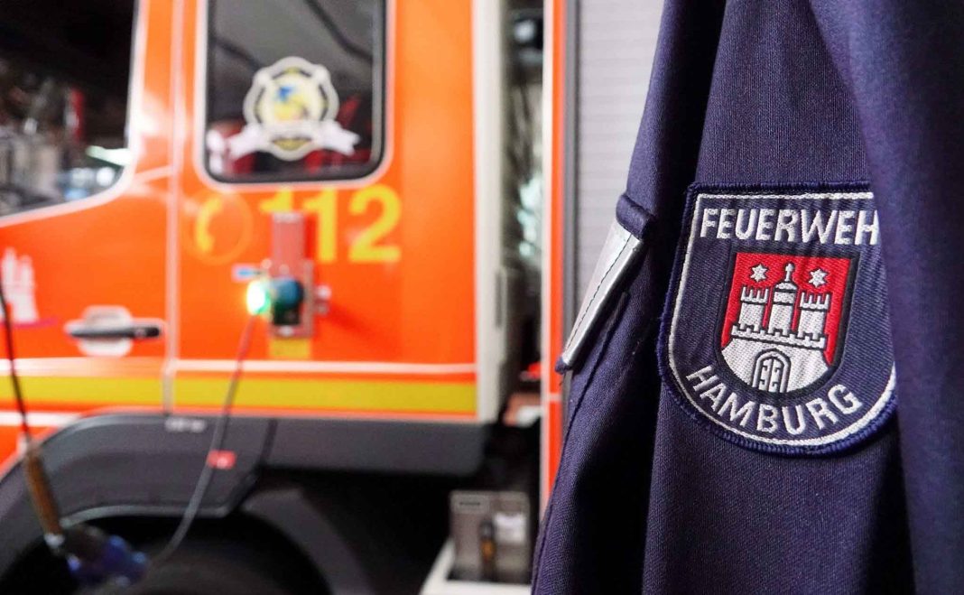 Im Hintergrund ist ein oranges Feuerwehrauto zu sehen. Rechts im Vordergrund sieht man eine blaue Jacke mit einem 