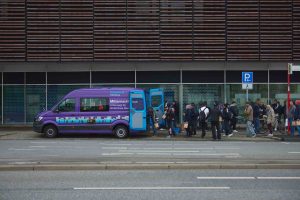 Links steht der lila/ blaue Mitternachtsbus vor einem Gebäude. Nach rechts weg steht eine Schlange obdachloser Menschen, die bis aus dem Bild geht. Sie stehen an, um Getränke und Wasser zu bekommen.