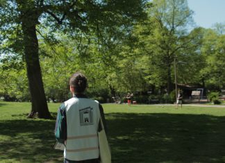 Streitfall Außenwerbung: Zwischen Litfaßsäule und LED-Screen Ein Mann im grünen Hemd, mit weißer Warnweste und Jutebeutel läuft durch einen Park. Im Hintergrund sind unscharf zwei Menschen zu sehen, die Federball spielen. Auf dem Rücken der Weste steht "Hamburg Werbefrei"