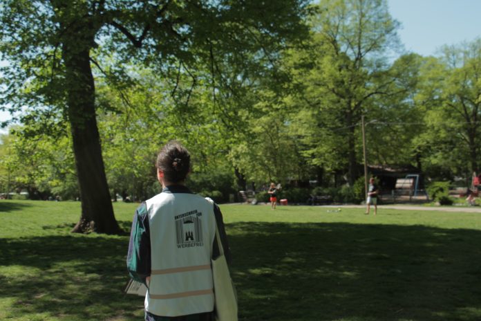 Ein Mann im grünen Hemd, mit weißer Warnweste und Jutebeutel läuft durch einen Park. Im Hintergrund sind unscharf zwei Menschen zu sehen, die Federball spielen. Auf dem Rücken der Weste steht 