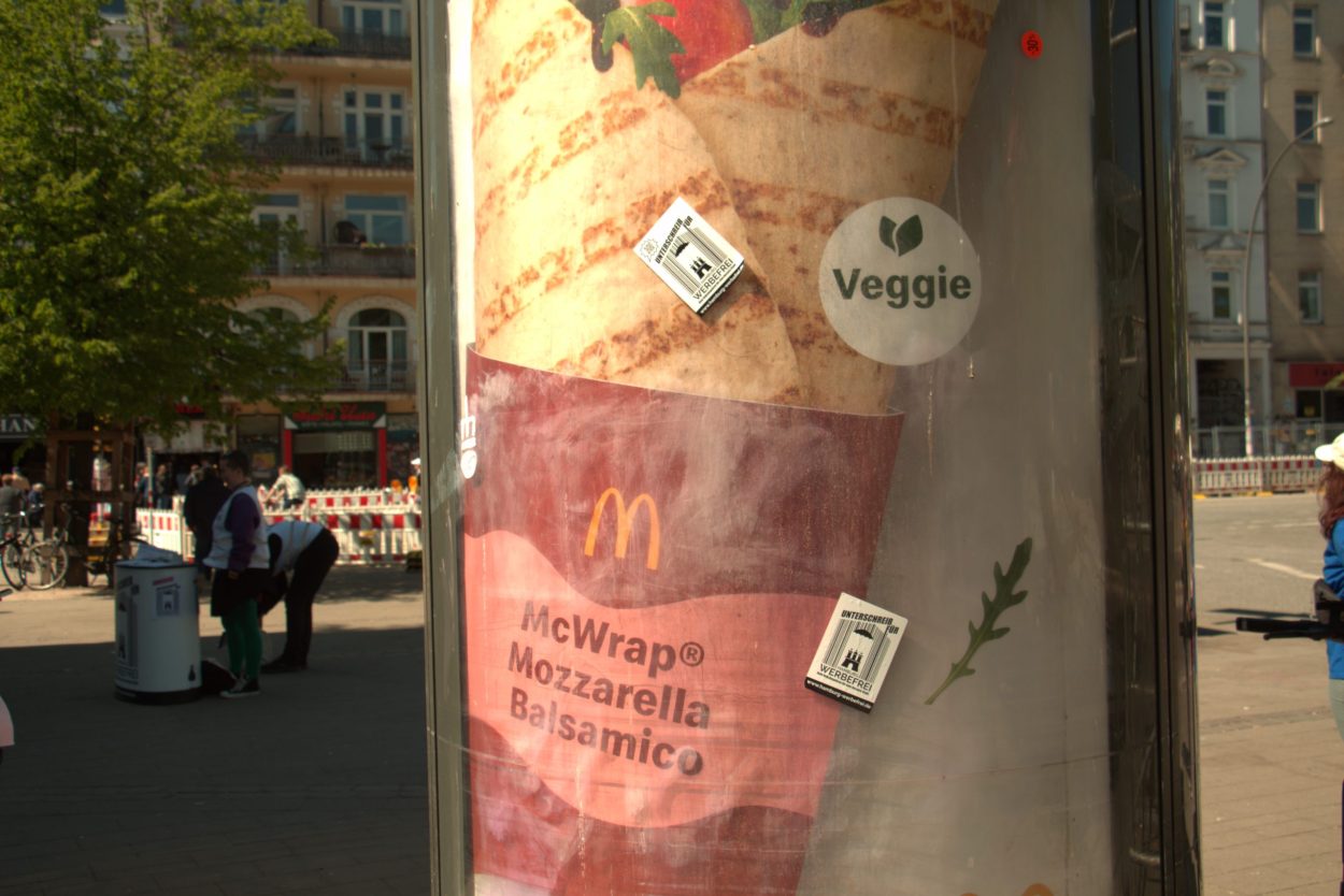 Eine Littfaßsäule mit einem Wrap von McDonalds darauf. Sie ist dreckig. Darauf kleben Sticker von "Hamburg Werbefrei". Im Hintergrund stehen unscharf zwei Menschen an einem Infostand der Initiative.