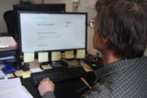 Ein Mann mit dunklen Haaren und Brille liest eine Website auf einem Computer. Er sitzt an seinem Schreibtisch im Schreibservice und ist Teilnehmender an den Arbeitsgelegenheiten.
