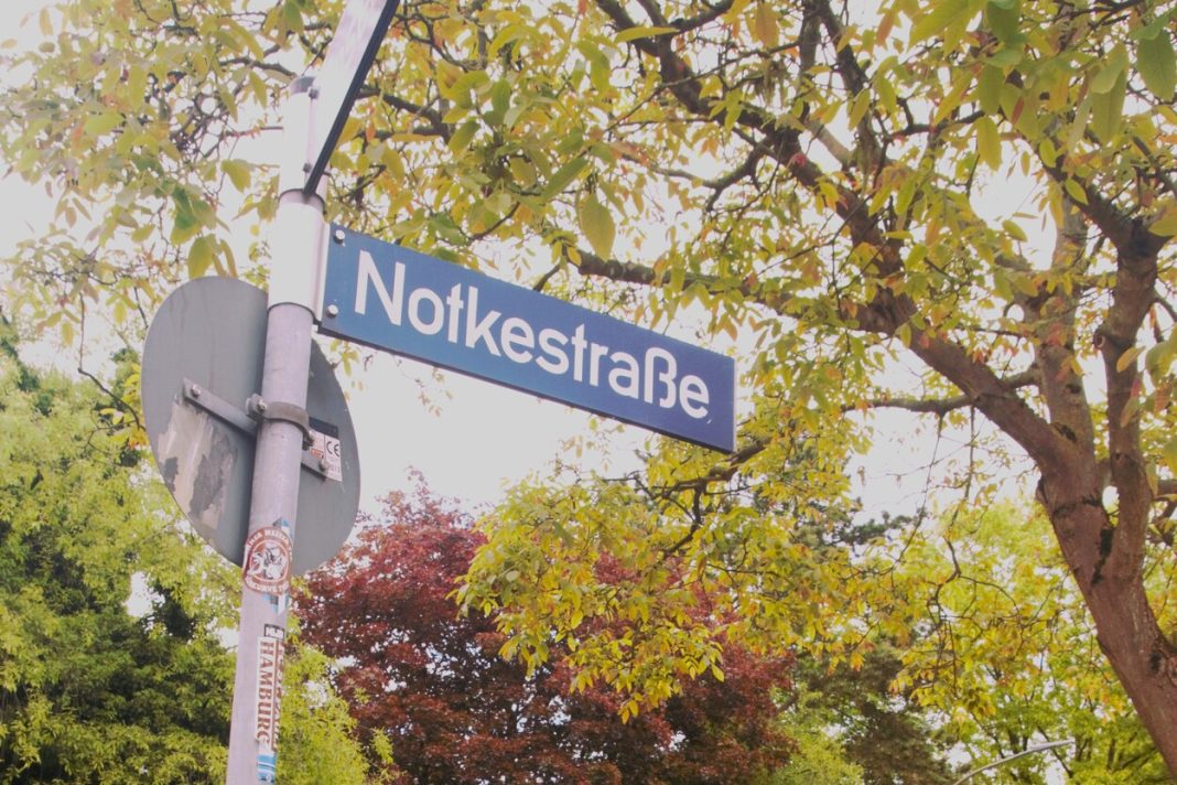 Ein Straßenschild, auf dem Notkestraße steht. Im Hintergrund sind Bäume.