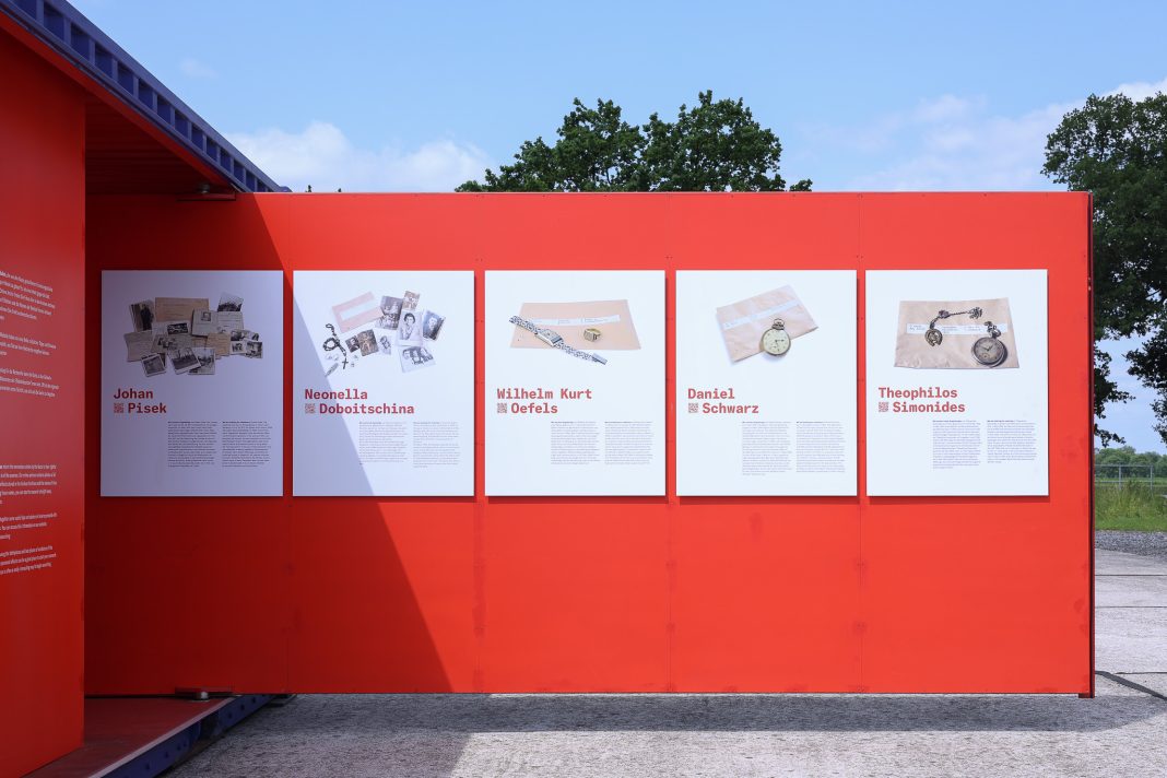 Eine rote Wand eines Ausstellungscontainers ragt auf den Platz des ehemaligen Konzentrationslagers in Neuengamme. Es sind mehrere Informationsplatten an der Wand befestigt. Sie zeigen Gegenstände von den Opfern der NS-Verbrechen.