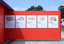 #StolenMemory – auf der Suche nach Angehörigen der NS-Opfer Eine rote Wand eines Ausstellungscontainers ragt auf den Platz des ehemaligen Konzentrationslagers in Neuengamme. Es sind mehrere Informationsplatten an der Wand befestigt. Sie zeigen Gegenstände von den Opfern der NS-Verbrechen.