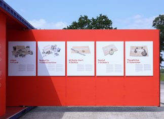 #StolenMemory – auf der Suche nach Angehörigen der NS-Opfer Eine rote Wand eines Ausstellungscontainers ragt auf den Platz des ehemaligen Konzentrationslagers in Neuengamme. Es sind mehrere Informationsplatten an der Wand befestigt. Sie zeigen Gegenstände von den Opfern der NS-Verbrechen.