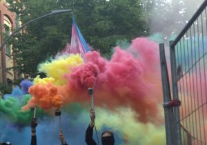 Zu sehen sind eine vermummte Person und die Hände drei anderer Demonstrierenden, die Bengalo Feuer mit verschiedenen farben in die Höhe halten.