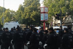 Das Bild zeigt mehrere Polizist*innen von hinten, als sie vor der Demonstration hergehen. Sie tragen dunkelblaue Uniformen und halten weiße Helme in der Hand.