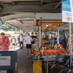 TikTok-Trend Wochenmarkt Der Markt mit Obstständen unter der U-Bahn Brücke. Einige Menschen laufen umher. Das Schild zeigt die Aufschrift Isemarkt.