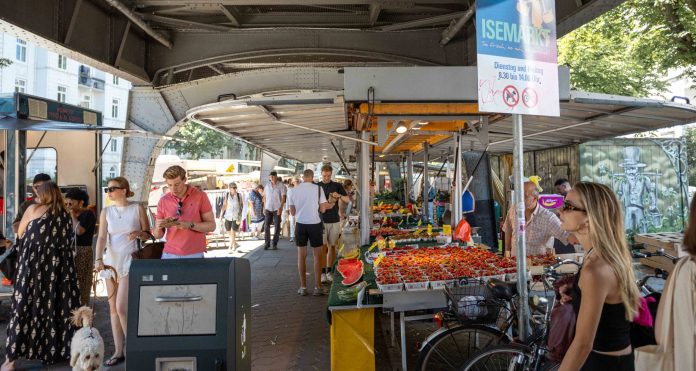 Der Markt mit Obstständen unter der U-Bahn Brücke. Einige Menschen laufen umher. Das Schild zeigt die Aufschrift Isemarkt.