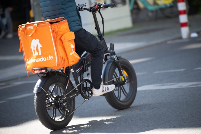 Lieferando Eine Person sitzt auf einem Fahrrad. Auf dem Gepäckträger ist eine orangefarbene Thermobox mit dem Lieferando-Logo befestigt.
