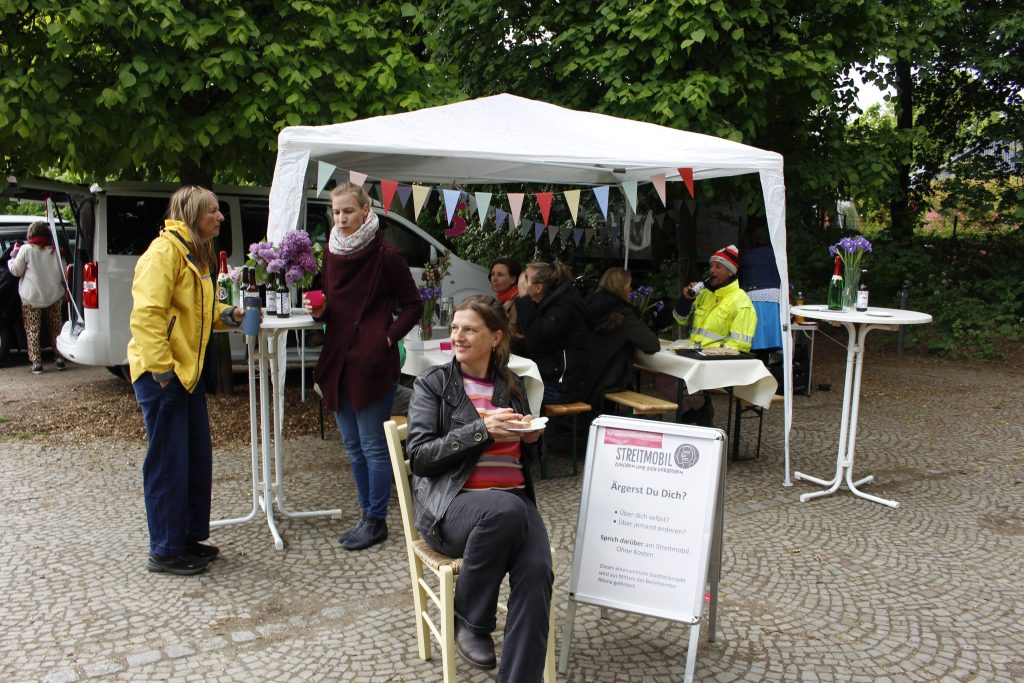 Katja Kaiser sietzt vor ihrem STreitmobil und einem Pavillion auf einem Stuhl mit einem Kuchen in der Hand.