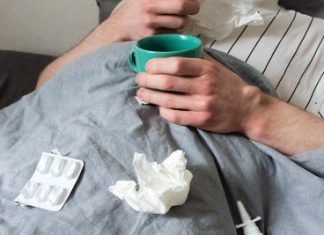Influenza auf dem Vormarsch: Deutlich mehr Tote und Infizierte Ein Mann liegt mit Taschentüchern, Teetasse, Nasenspray und Tabletten im Bett und symbolisiert die gestiegenen Influenza zahlen.