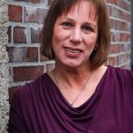 Sexualbegleiterin Bianka Pille Ein Porträt von Bianka Pille. Sie lehnt an einer Backsteinwand und schaut lächelnd in die Kamera. Sie trägt ein weinrotes Oberteil.