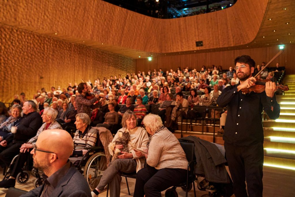 Mehrere ältere Menschen, sowohl Männer als auch Frauen, sitzen in einem Konzertsaal. Rechts steht ein Mann. Er trägt ein schwarzes Hemd und eine schwarze Hose. Er spielt eine Violine.