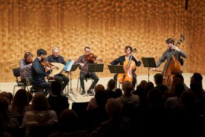 Fünf Männer und eine Frau sitzen auf Stühlen auf der Bühne. Sie spielen verschiedene Instrumente. Vorne ist ein Publikum von Hinten zu sehen. 