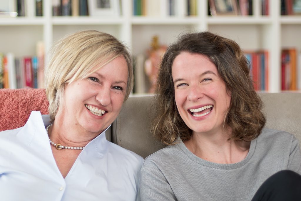 Zwei lachende Frauen sitzen auf einem Sofa vor einem Bücherregal.