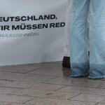 Müssen wir reden? Die Hosenbeine einer Frau in Jeans vor einem Tisch mit einem Banner mit der Aufschrift "Deutschland wir müssen reden". Sie steht vor einem Schwarz-Weiß-Foto von Charlie Kirk und der Boden ist nass.