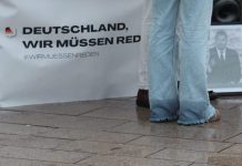 Müssen wir reden? Die Hosenbeine einer Frau in Jeans vor einem Tisch mit einem Banner mit der Aufschrift "Deutschland wir müssen reden". Sie steht vor einem Schwarz-Weiß-Foto von Charlie Kirk und der Boden ist nass.