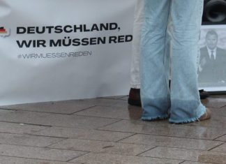 Müssen wir reden? Die Hosenbeine einer Frau in Jeans vor einem Tisch mit einem Banner mit der Aufschrift "Deutschland wir müssen reden". Sie steht vor einem Schwarz-Weiß-Foto von Charlie Kirk und der Boden ist nass.