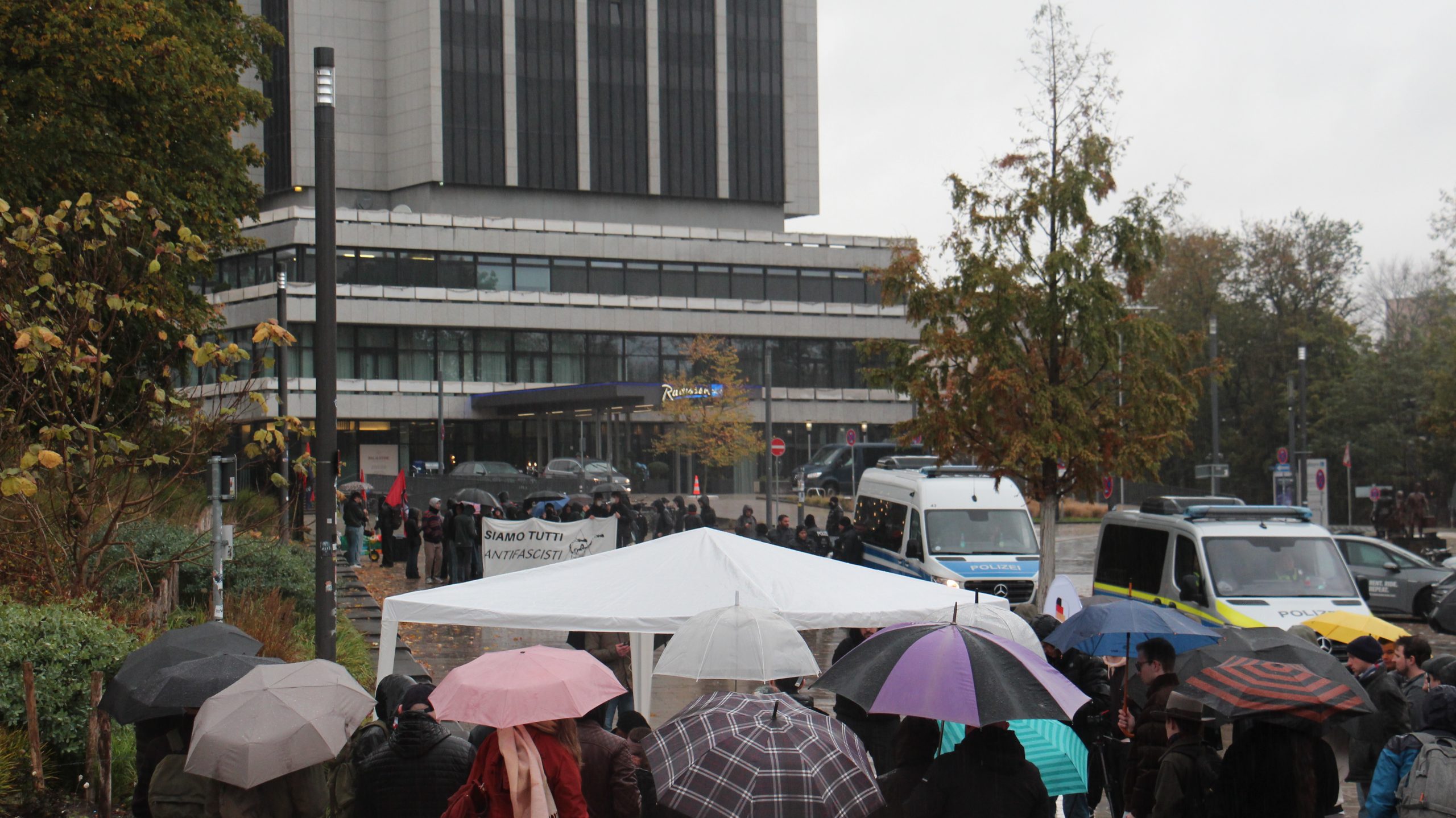 Zwei Demos stehen sich gegenüber. Im Hintergrund sieht man ein Banner "Siamo tutti antifascisti". Im Vordergrund sind viele Regenschirme und ein weißes Zelt zu sehen.