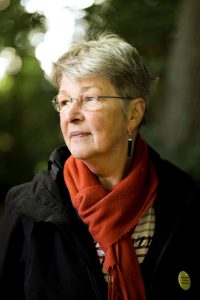 Ein Portrait von einer Frau. Der Hintergrund ist verschwommen, sie steht in einem Wald . Sie hat graue, kurze Haare und trägt eine Brille. Außerdem trägt sie einen orangefarbenen Schal.