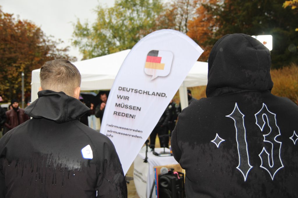 Zwei Menschen stehen im Regen mit dem Rücken zur Kamera vor einer Fahne mit dem Aufdruck "Deutschland, wir müssen reden!"