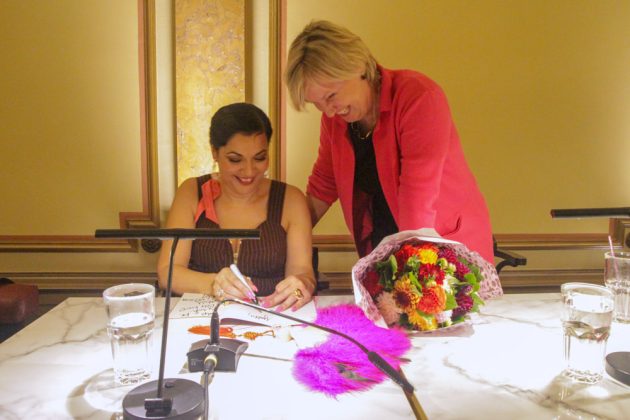 Zwei Frauen schauen lächeld auf ein Buch. Auf dem Tisch liegen Blumen.