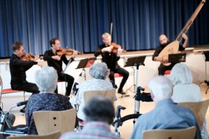 Vier Männer sitzen auf Stüheln und spielen verschiedene Instrumente. Sie tragen schwarze Kleidung. Vorne sitzen ältere Männer und Frauen. Sie sind von hinten zu sehen.