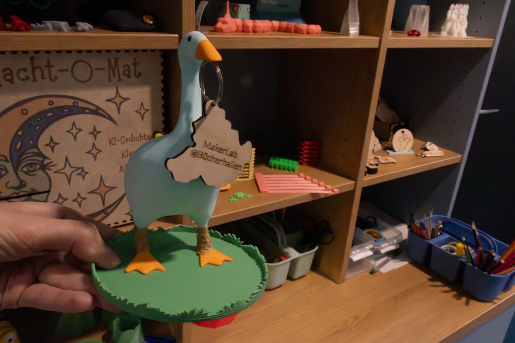 Eine 3D gedruckte hellblaue Gans mit orangenen Füßen hat einen Holzanhänger im Mund. Auf ihm steht MakerLab Bücherhalle.