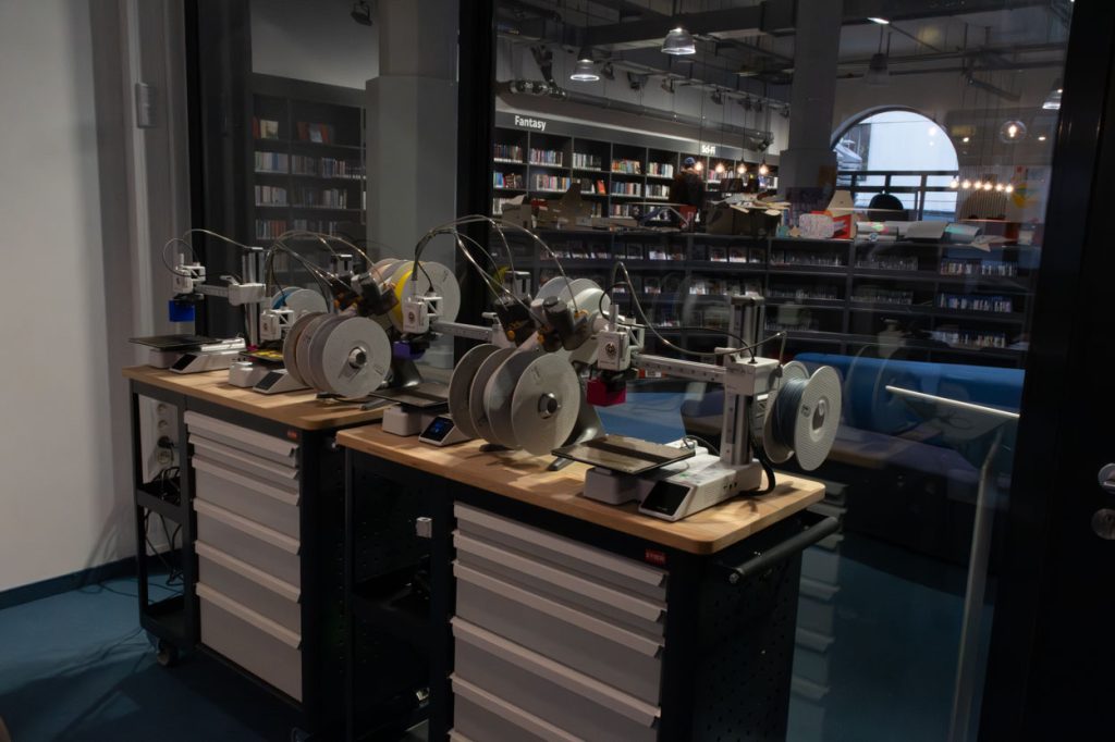 Zwei 3D-Drucker stehen auf zwei Rollwägen vor einer Glaswand.