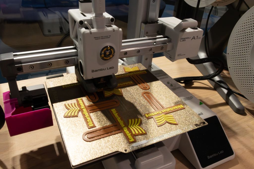 Der Druckarm eines 3D-Druckers fährt über eine Druckplatte. Auf der Druckplatte liegen fertig gedruckte Lesezeichen der Bücherhalle.