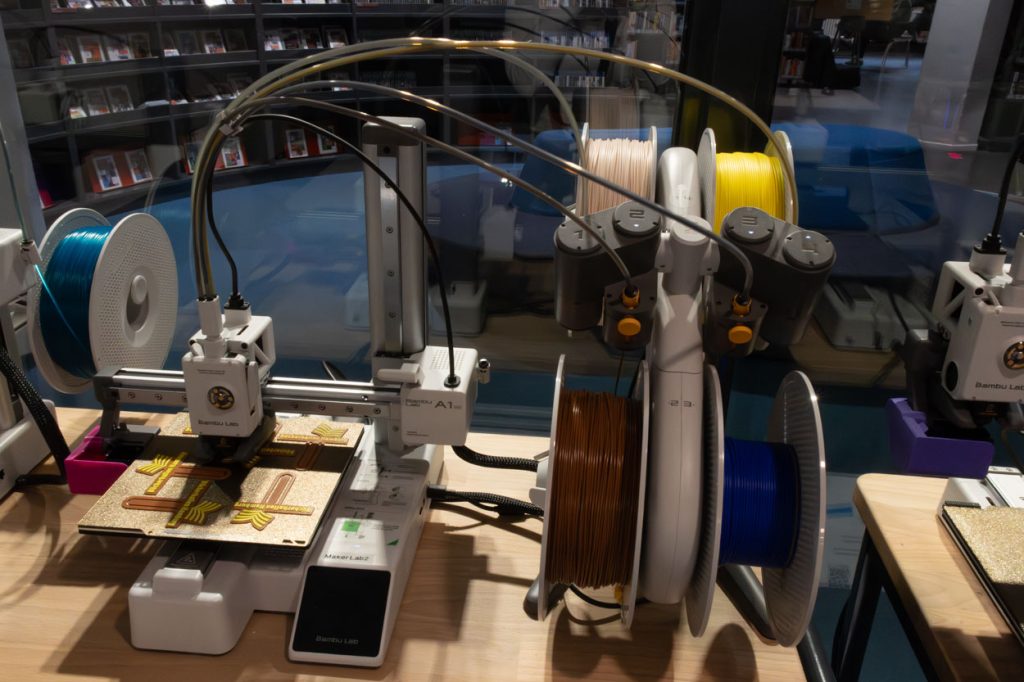 Ein 3D-Drucker steht vor einer Glaswand. Am Ende des 3D-Druckers sind vier Spulen angebracht. Auf ihnen ist das Druckmaterial, Filament aus PLA, in verschiedenen Farben aufgewickelt.