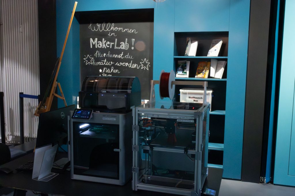 Ein 3D-Drucker steht vor einer blauen Wand. Auf einer Tafel an der blauen Wand steht Wilkommen im MakerLab. Hier kannst du kreativ werden.
