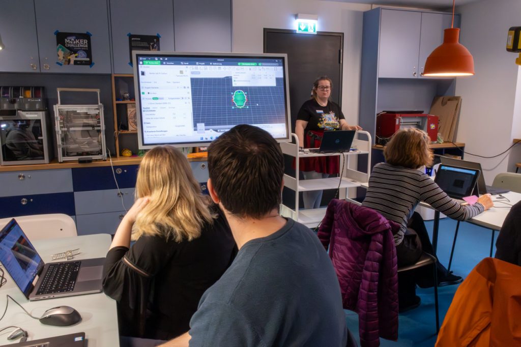 Eine Frau steht neben einem großen Bildschirm und erklärt ein 3D-Druck-Programm. Vor ihr sitzen drei Personen an Tischen in einem Raum.