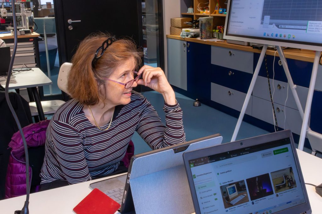 Eine Frau mit braunen kurzen Haaren rückt ihre rosane Brille auf der Nase zurecht. Sie sitzt vor einem Laptop und hört gespannt einem Vortrag zu.
