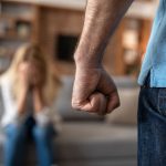 Leichter Ausstieg aus Mietverträgen für Gewaltopfer Rechts im Vordergund ist die Faust und der Körper eines Mannes abgebildet. Im Hindergrund sitzt eine Frau auf dem Sofa. Sie hat blonde Haare und die Arme vor ihrem Gesicht verschränkt.