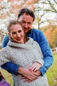 Das Bild zeigt einen Mann in einem blauen Pullover und mit dunklen Haaren, der eine Frau mit blonden Haaren und in grauem Pullover von hinten umarmt. Beide lächeln in die Kamera. Im Hintergrund sieht man eine Wiese und einen herbstlichen Baum.