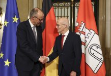 Merz zu Besuch in Hamburg Der Bundeskanler Friedrich Merz und der Bürgermeister Peter Tschentscher recihen sich die Hände. Im Hintergrund sind Flaggen aufgestellt.