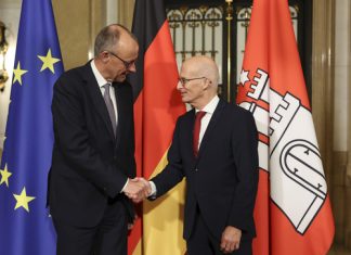 Merz zu Besuch in Hamburg Der Bundeskanler Friedrich Merz und der Bürgermeister Peter Tschentscher recihen sich die Hände. Im Hintergrund sind Flaggen aufgestellt.