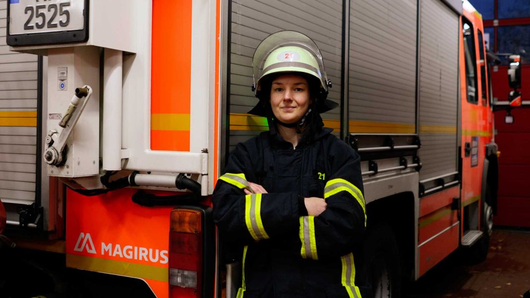 Nora Tenbusch-1 Feuerwehrfrau Nora Tenbusch steht vor einem Einsatzwagen der Feuerwehr. Sie trägt die Schutzkleidung der Feuerwehr und hat einen Helm auf dem kopf. Sie schaut in die Kamera und hat ihre Arme verschränkt.