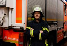 Feuerwehrfrau Nora Tenbusch Feuerwehrfrau Nora Tenbusch steht vor einem Einsatzwagen der Feuerwehr. Sie trägt die Schutzkleidung der Feuerwehr und hat einen Helm auf dem kopf. Sie schaut in die Kamera und hat ihre Arme verschränkt.