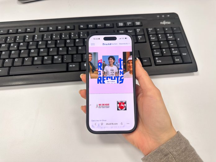 Onlineshop Druck18 Man sieht bei dem Blick über die Schulter einer Person den geöffneten Onlineshop Druck18.com auf einem Handy, vor einem Schreibtischhintergrund.